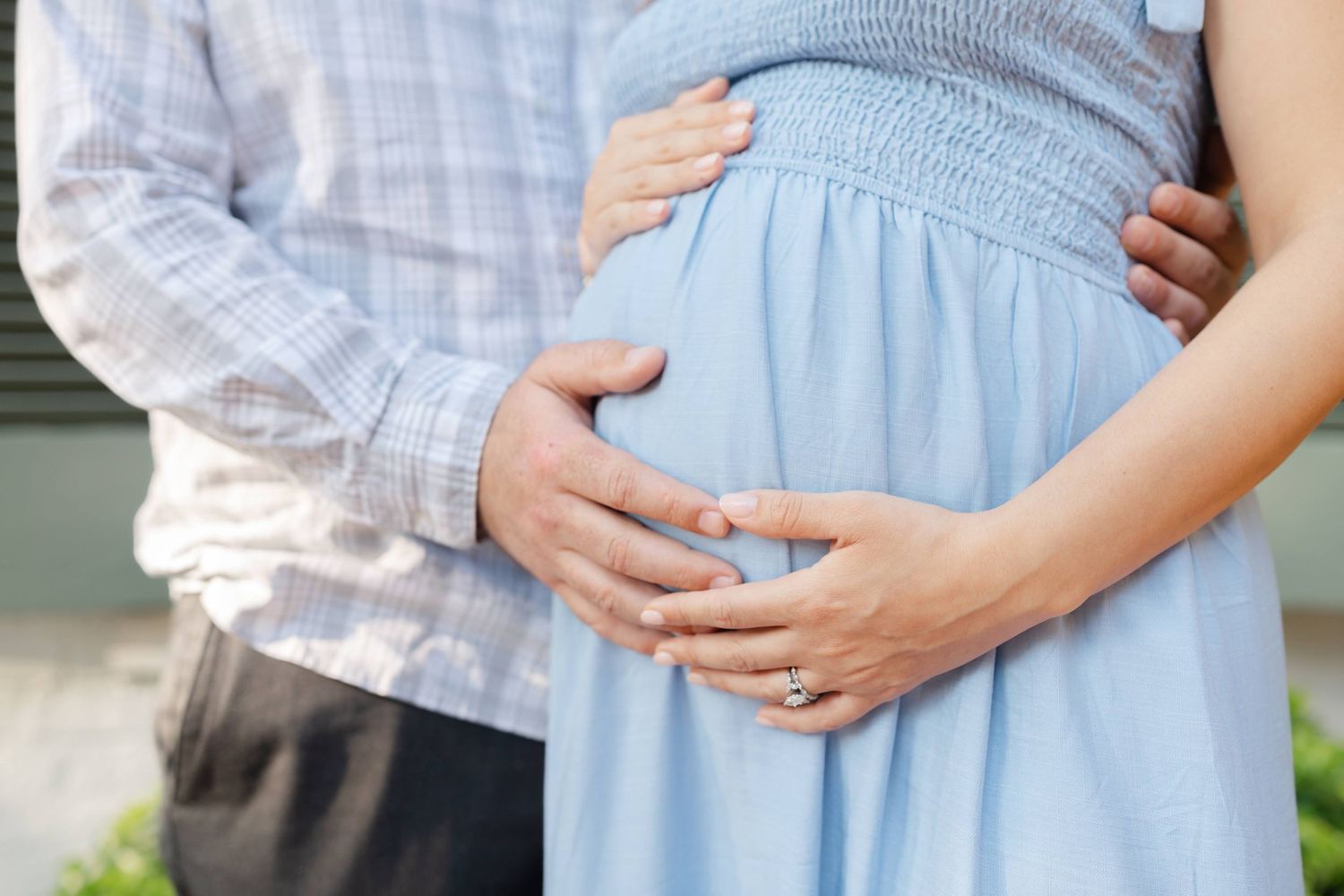 Couple tenderly cradling pregnant belly outdoors, man in plaid shirt embracing woman in light blue maternity dress.