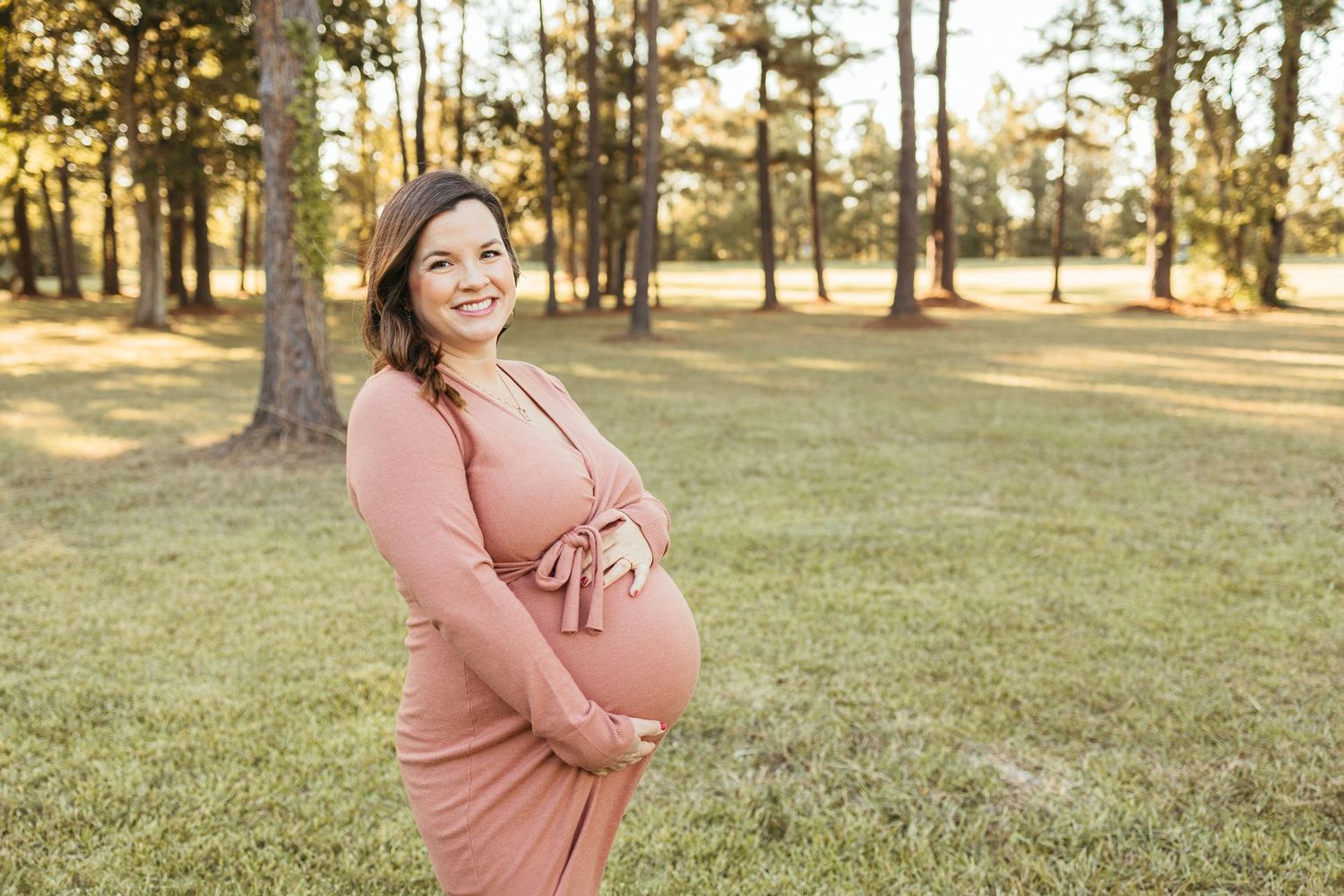 A pregnant woman in a pink dress smiles while cradling her baby bump in a sunny outdoor park.