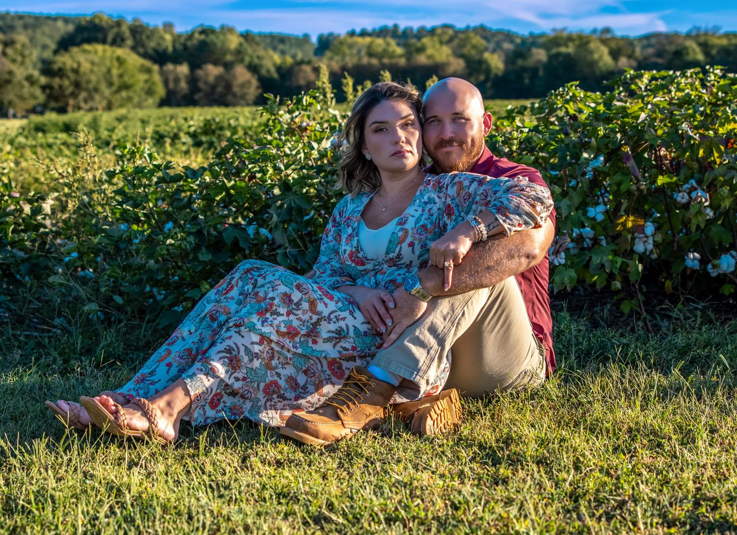 Couple sitting together on grass in a vineyard, woman in floral dress leaning on man in red shirt.