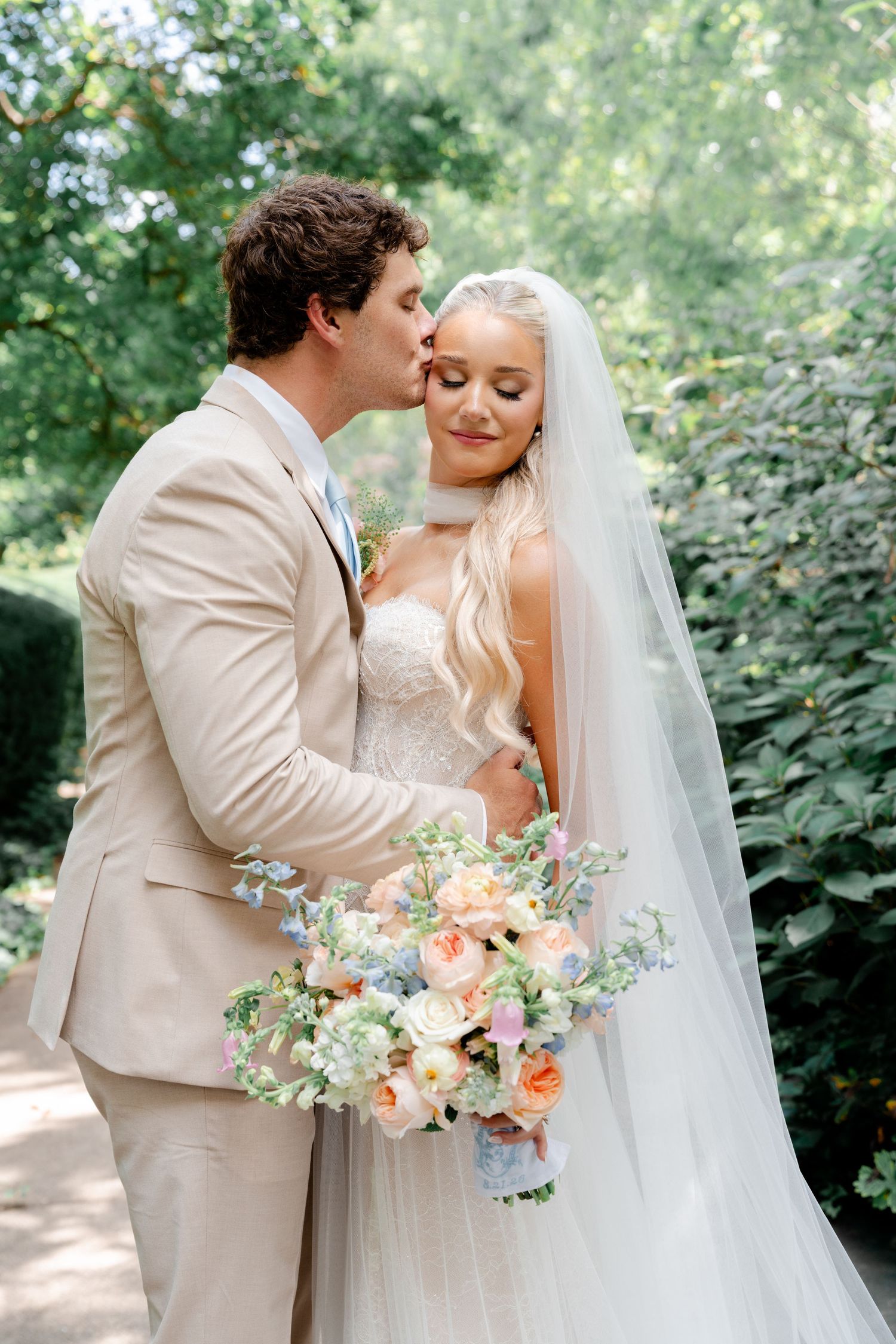 Groom in tan suit kisses bride wearing white lace gown and veil, holding pastel floral bouquet in garden setting.