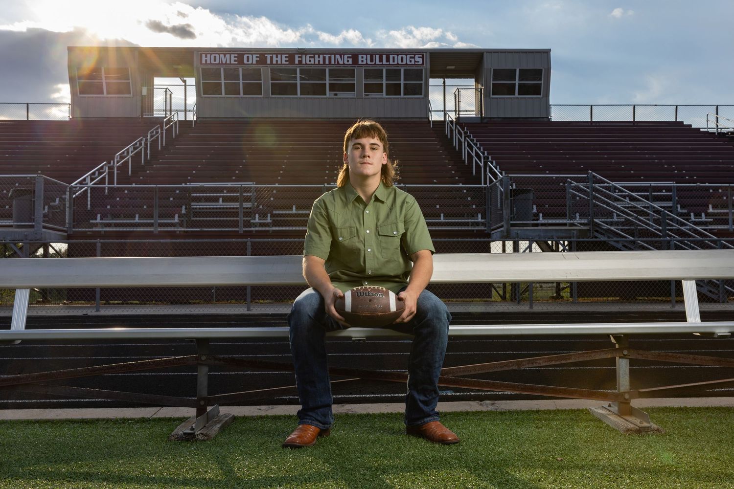 high school senior boy sitting on a bench at the stadium holding a football.