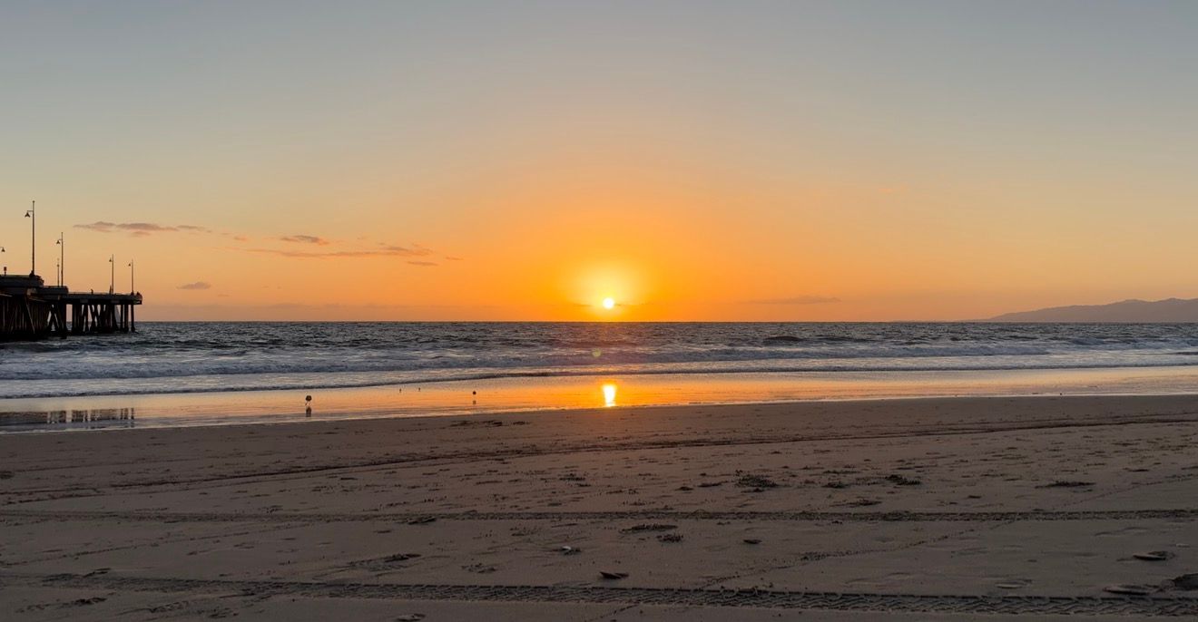 Golden sunset over calm ocean waters with a wooden pier silhouetted on the left and wet sandy beach in foreground.