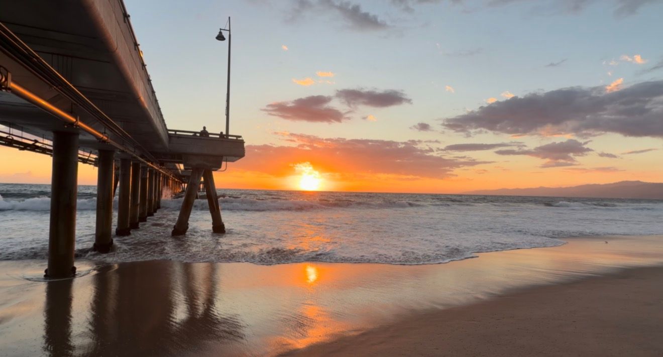 Golden sunset over the ocean horizon beside a wooden pier, with waves washing onto a wet sandy beach and reflecting warm orange sky colors.