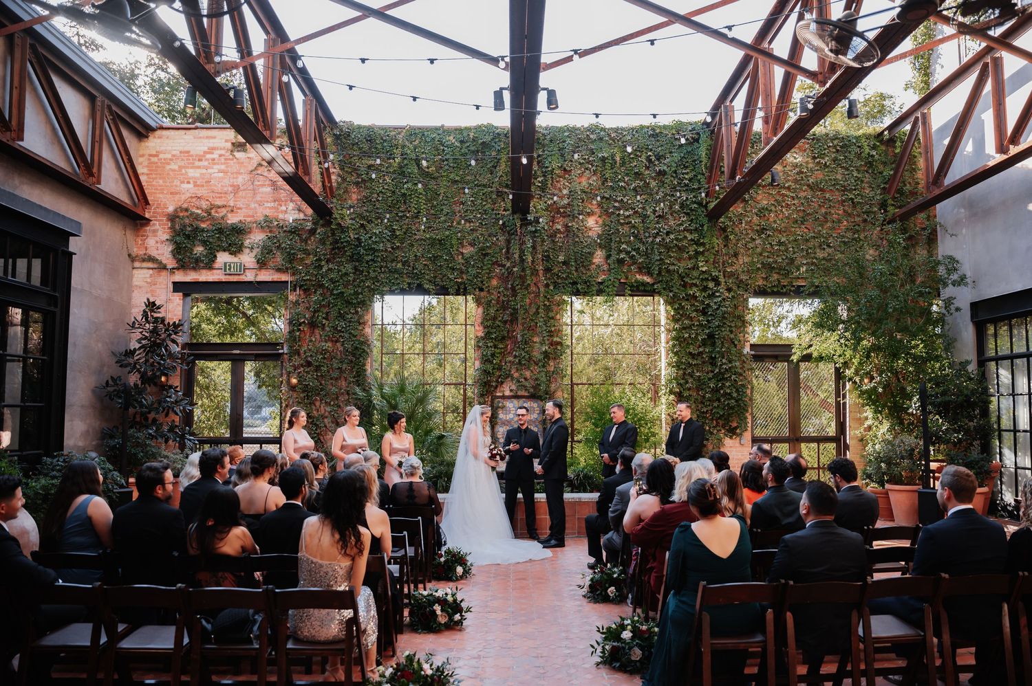 Bride and groom exchange vows at rustic indoor wedding ceremony with white chairs and floral arrangements.