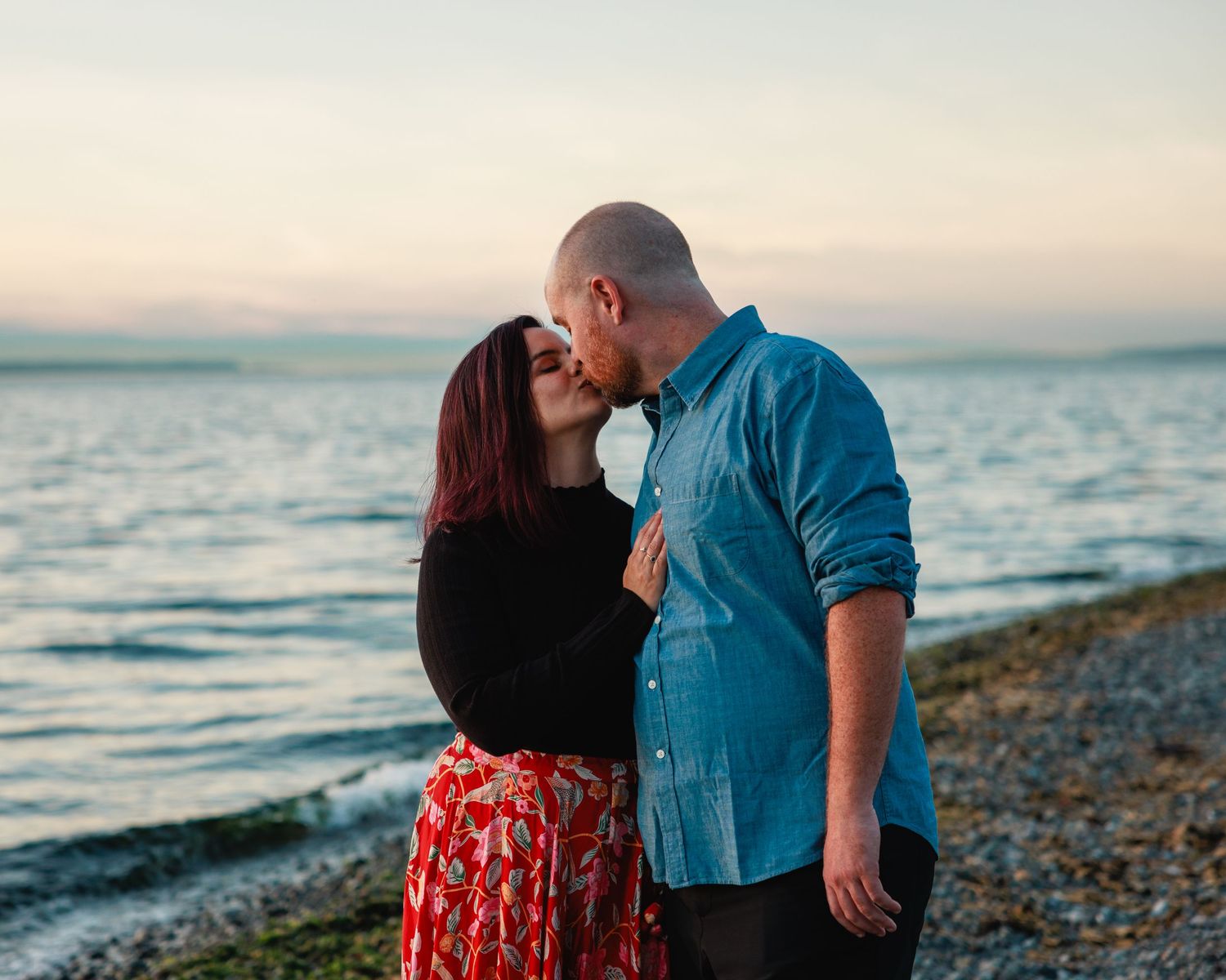 Romantic couple sharing a kiss on a pebble beach at sunset with calm ocean waters behind them.