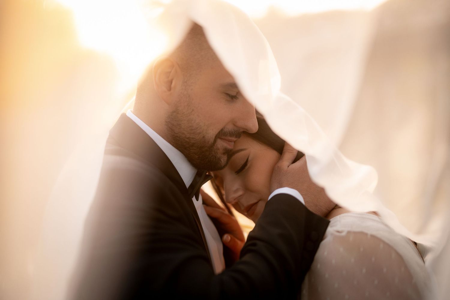 Newlywed couple shares an intimate moment under a veil during golden hour.