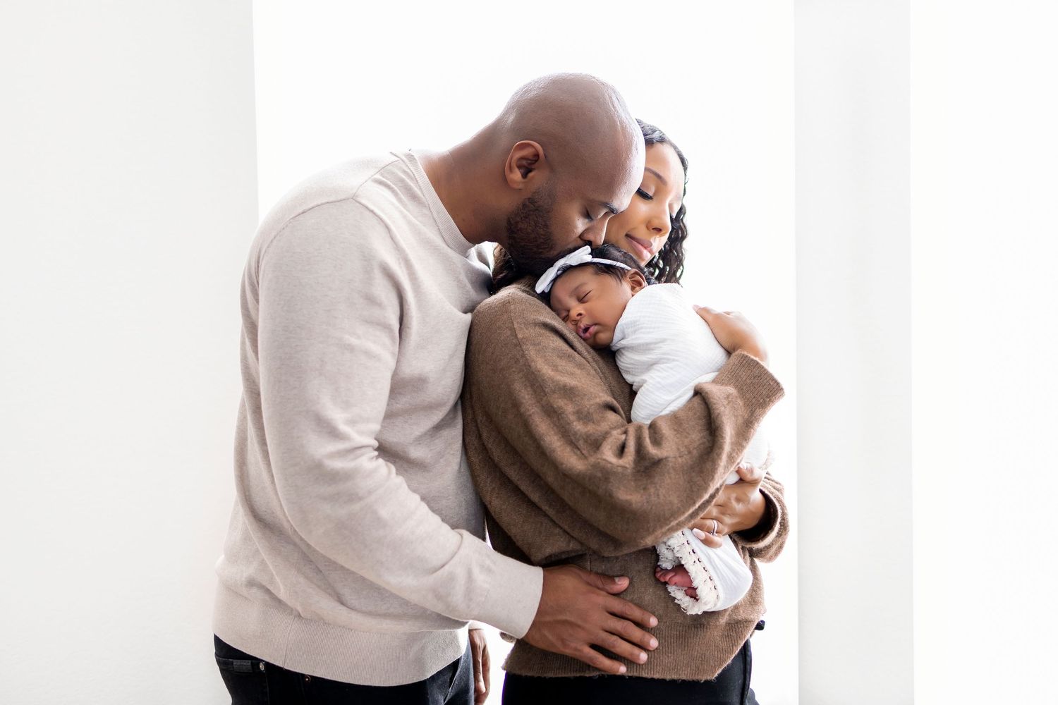 Family photo - Woman holding newborn baby girl on her chest, man behind her kissing baby's head, hand on womans hip