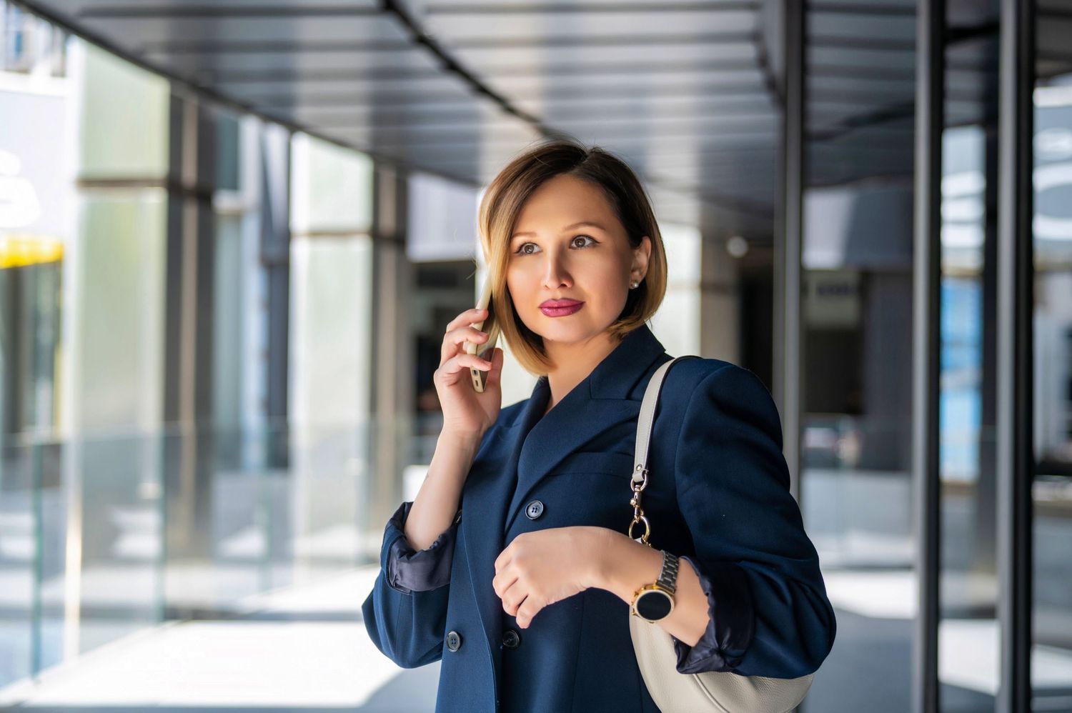 A person in a business suit talking on a mobile phone while standing in a modern office building corridor.
