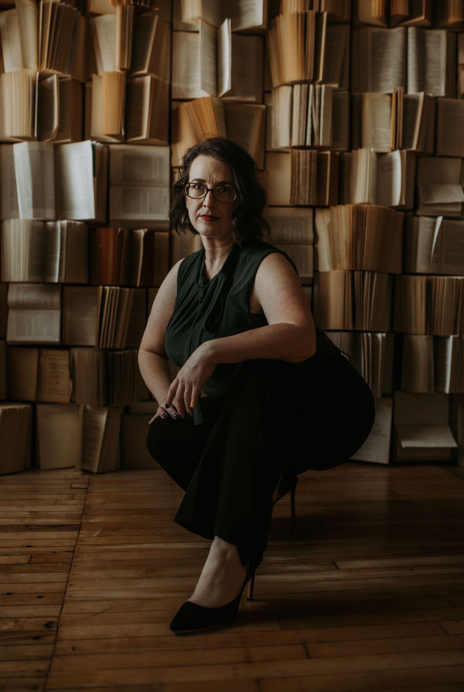 Artistic portrait against wall of stacked books in moody lighting.