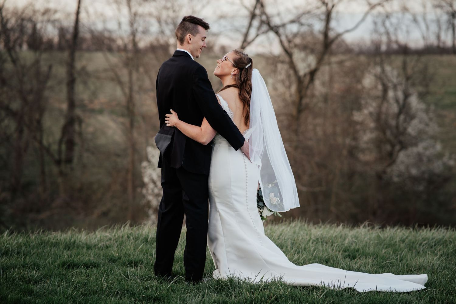 A wedding couple embraces on a grassy hilltop during golden hour.