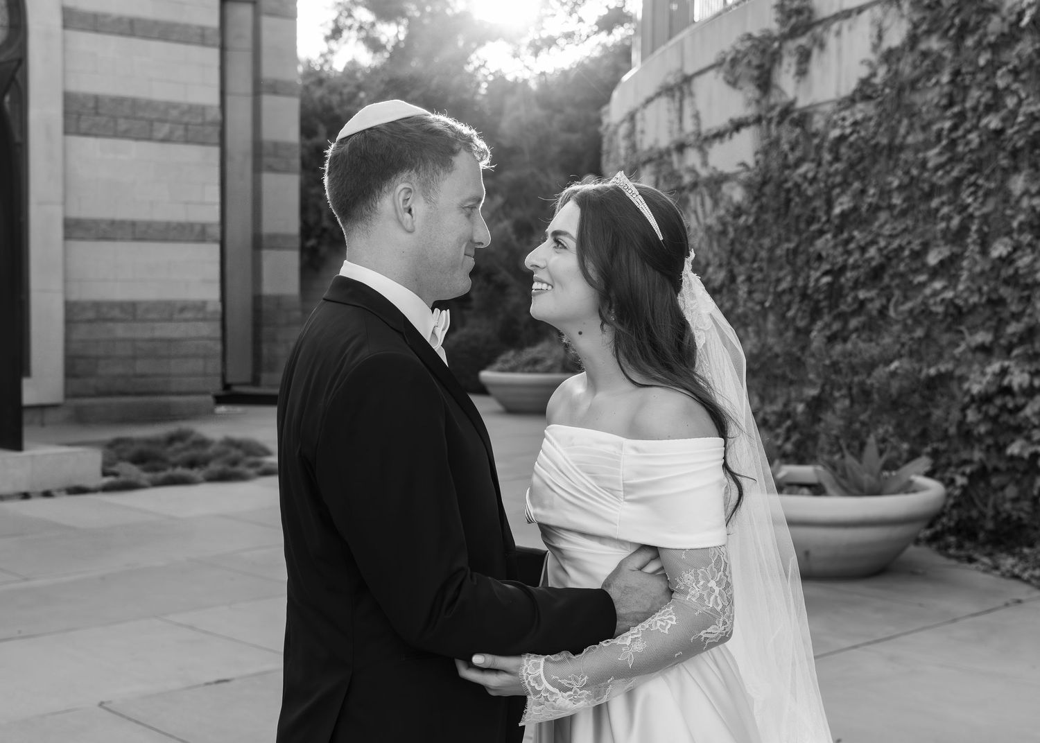Couple in formal wedding attire poses together in elegant outdoor setting with white columns at sunset.
