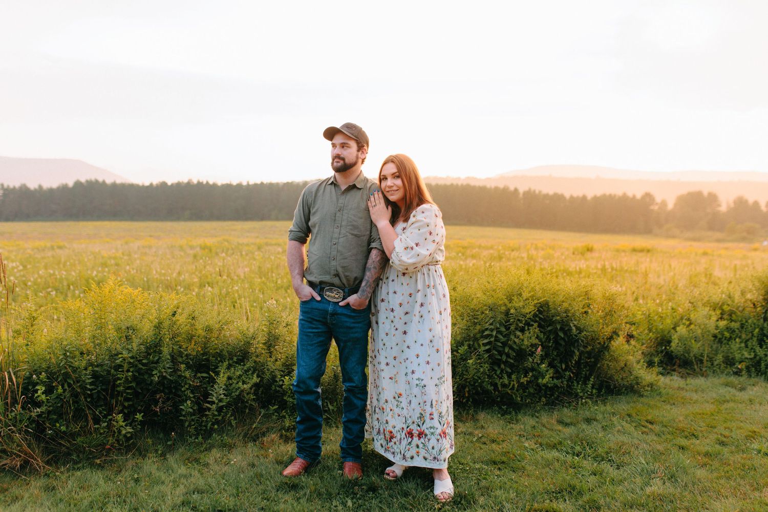 A couple stands together in a sunlit meadow during golden hour, with the woman wearing a floral dress.