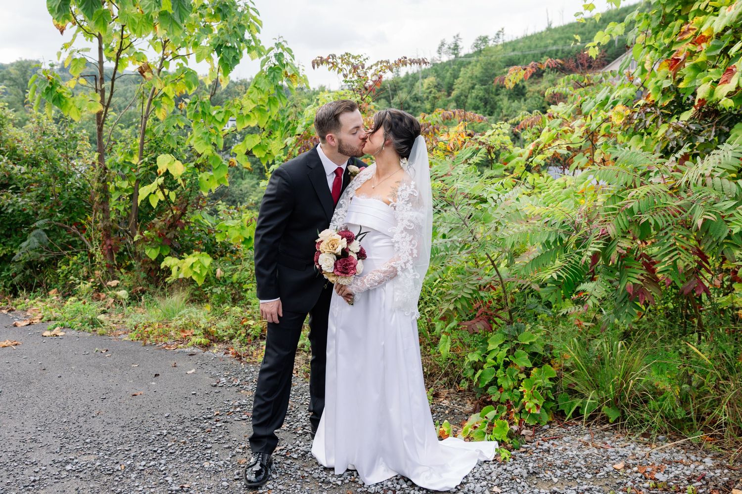 A couple in wedding attire shares an intimate moment on a gravel path surrounded by lush green foliage.