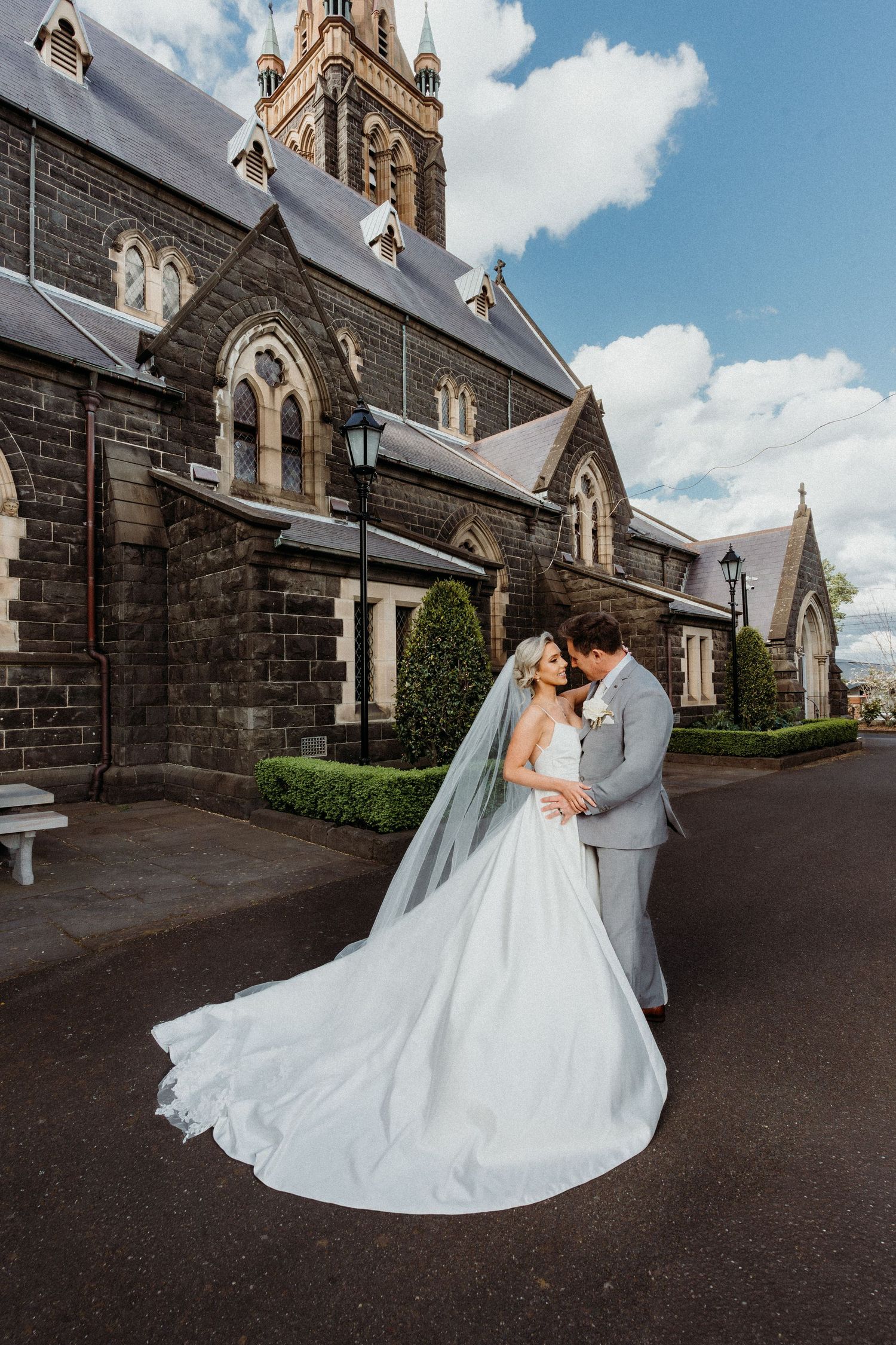 A romantic wedding couple embraces outside a gothic stone church with dramatic architecture and cloudy sky.