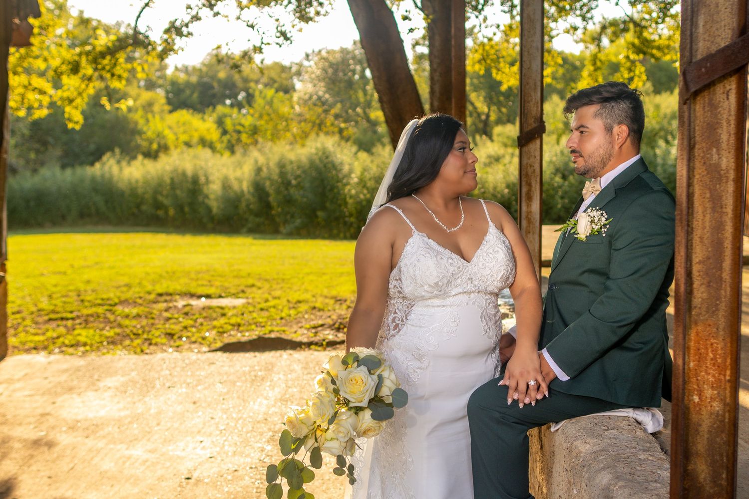 A couple in wedding attire sits together at sunset in a rustic outdoor setting with golden sunlight filtering through trees.