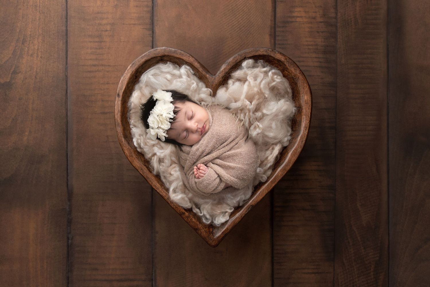 Baby sleeping peacefully in heart-shaped wooden bowl filled with soft white fabric.