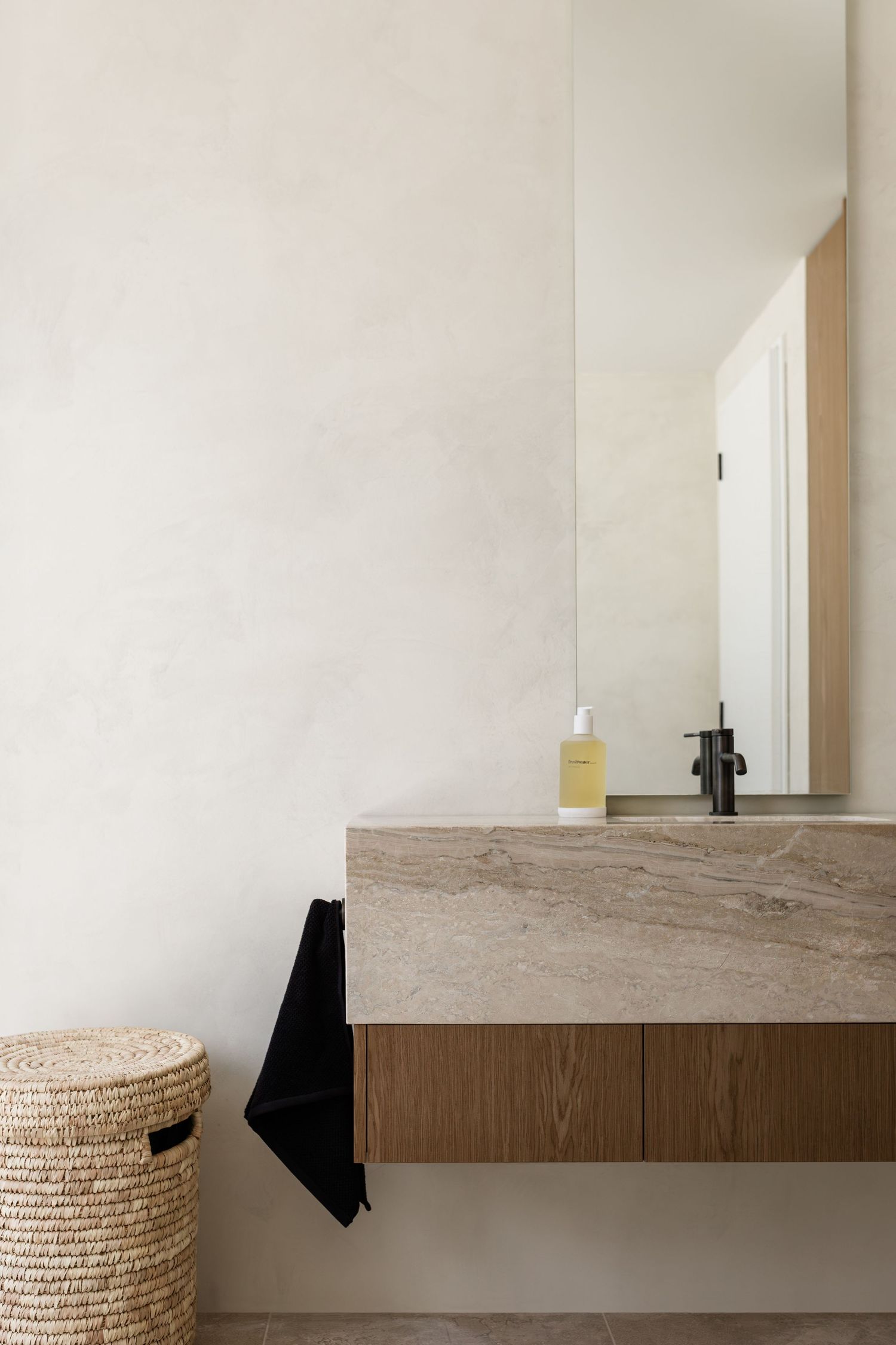 Minimalist white bathroom interior with wooden vanity, woven straw stools, and black towels against clean walls.