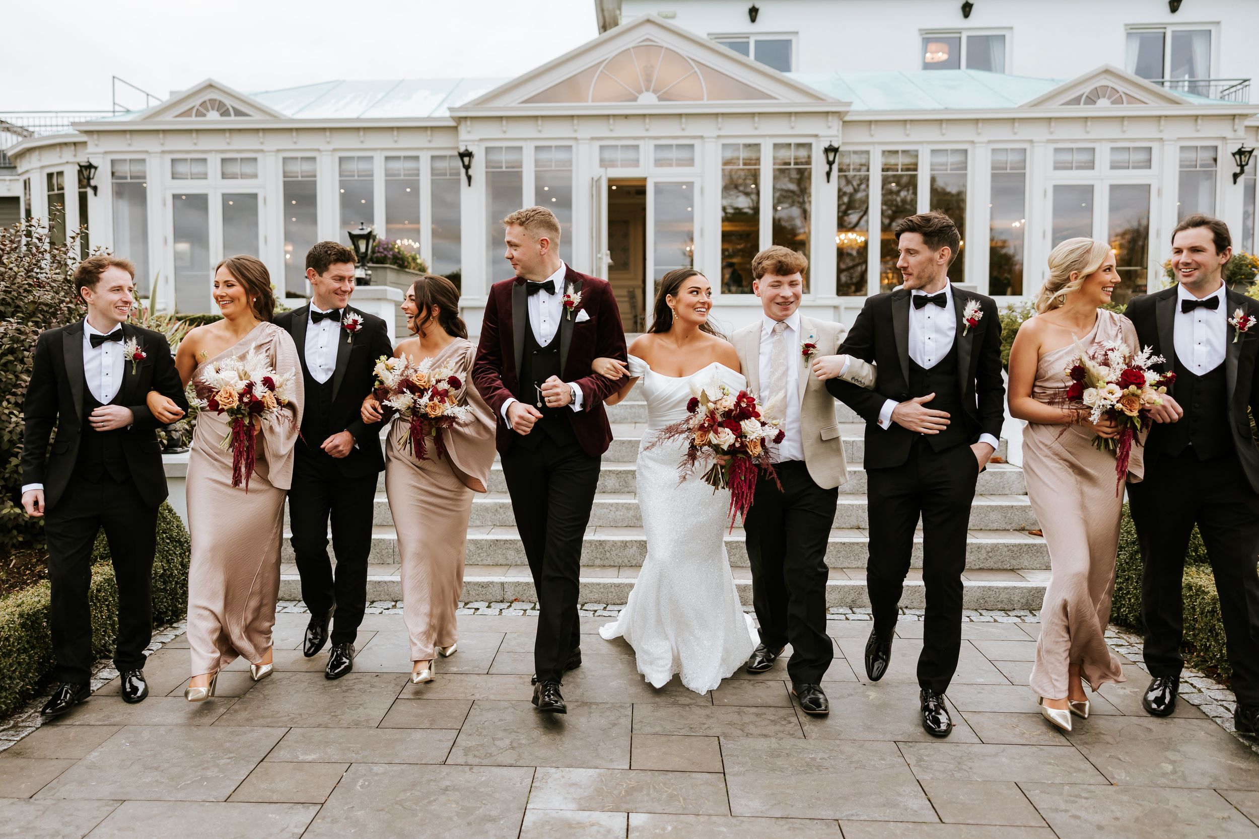 Wedding party in formal attire walks together and laughs on stone patio with white building in background.