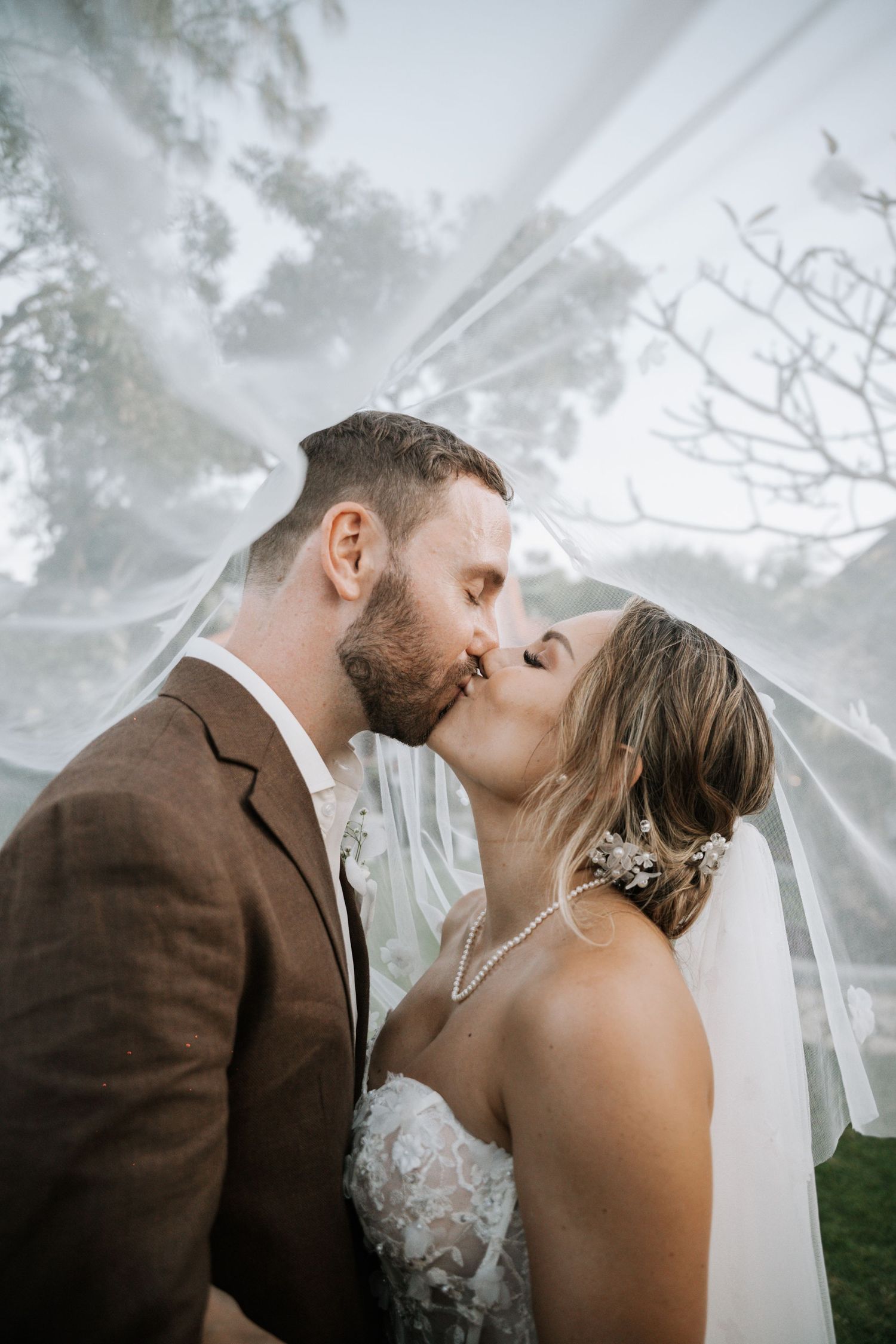 Silhouetted couple sharing a romantic kiss under flowing wedding veil at sunset with dramatic sky in background.