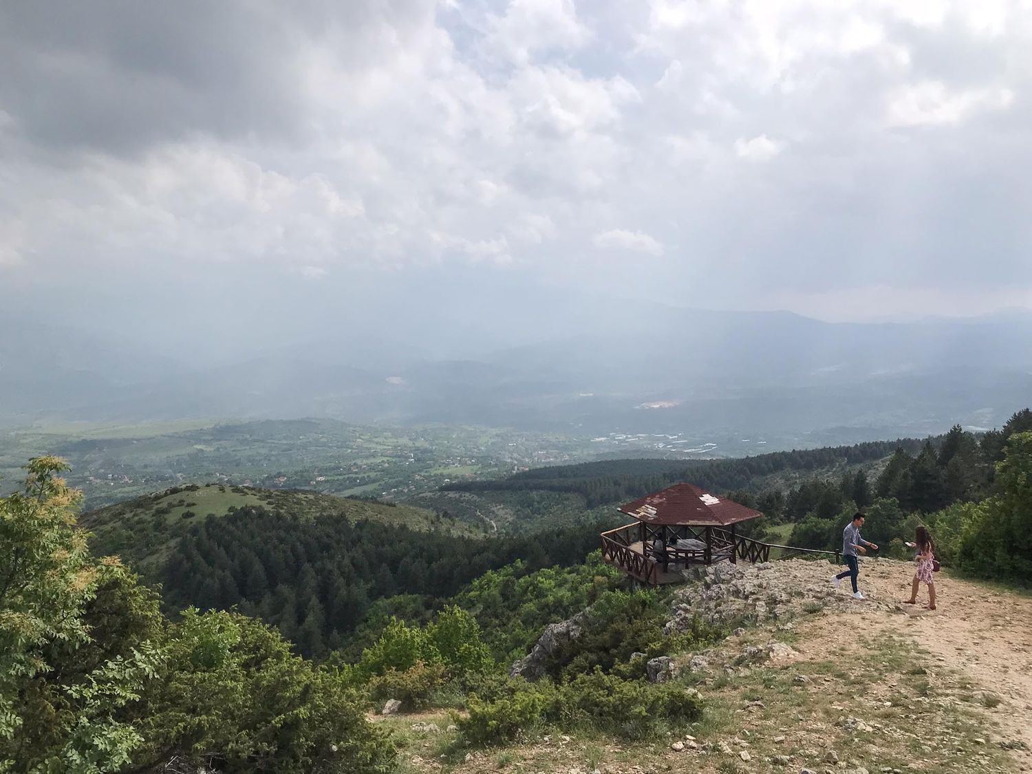 A scenic mountain viewpoint with a wooden gazebo overlooking green valleys and distant hills under cloudy skies.