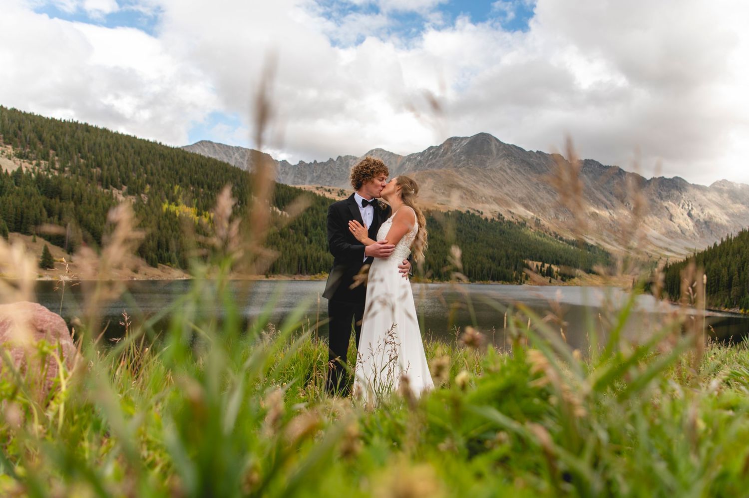 A couple in wedding attire stands on a mountainside slope with evergreen trees and dramatic mountain peaks in the background.