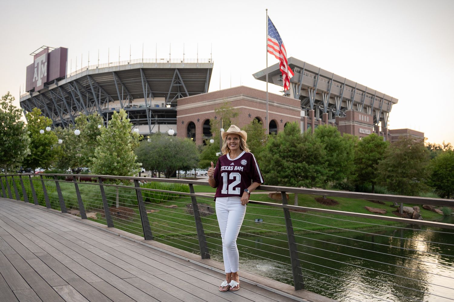Person wearing white sports jersey stands on wooden boardwalk along waterfront near urban buildings at sunset.