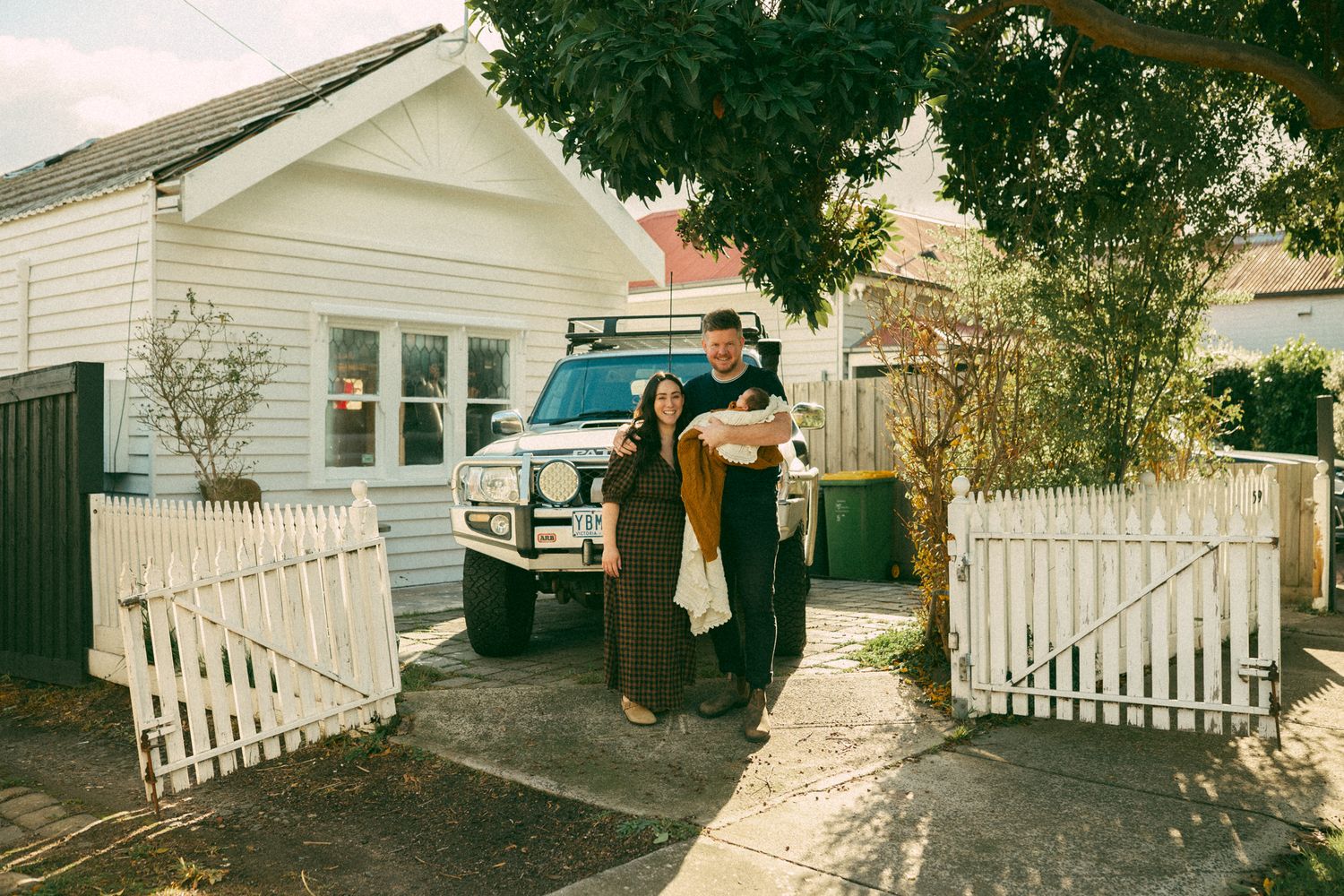 Two people embrace by a white picket fence with an ambulance parked in the driveway.