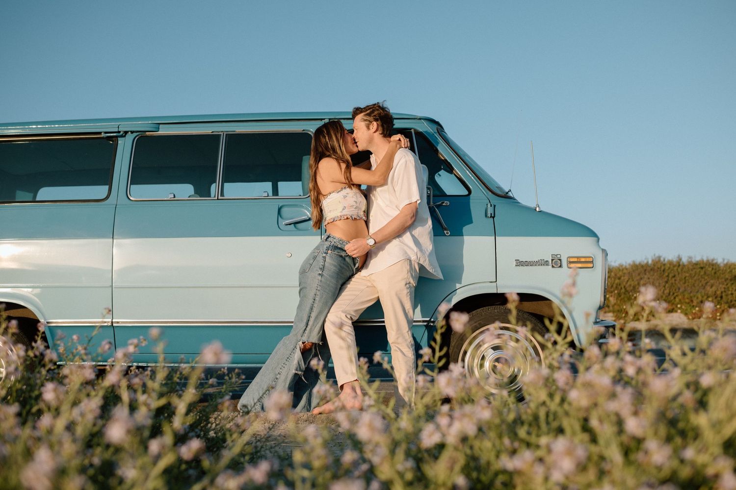 A romantic couple embraces next to a vintage blue van parked in a wildflower field during golden hour.