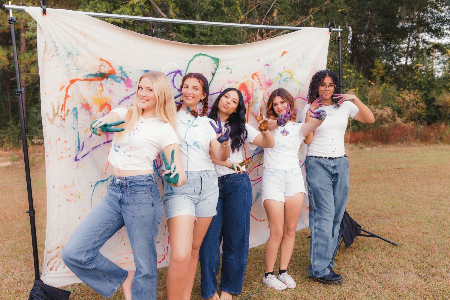 A group of friends in denim and white outfits pose together against a colorful painted backdrop outdoors.