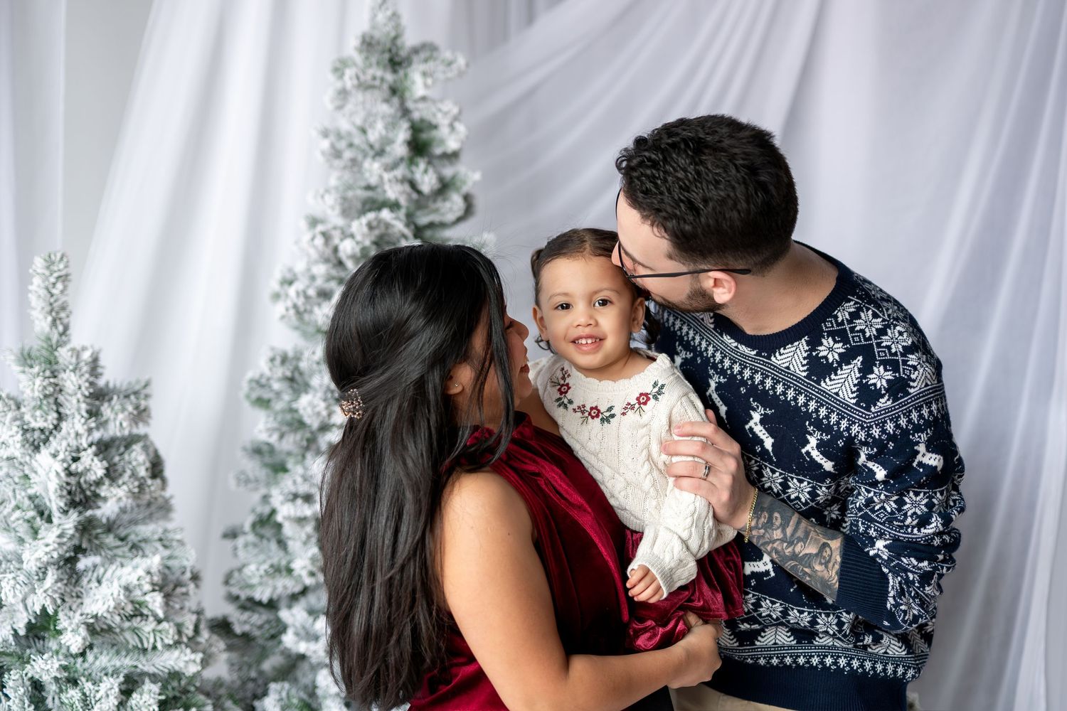 Parents in holiday sweaters kissing their toddler daughter near a snow-flocked Christmas tree.