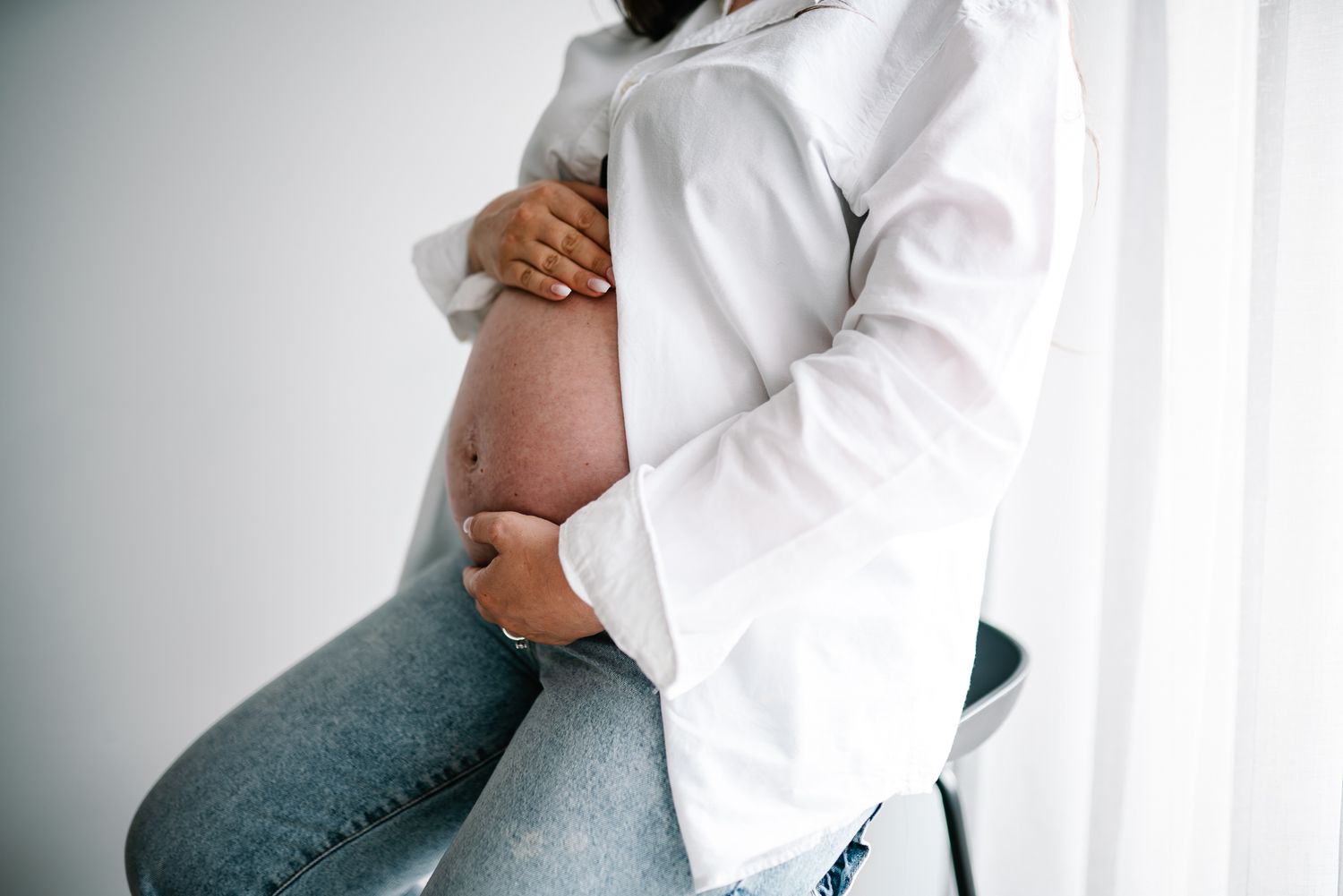 A pregnant belly is gently cradled while wearing a white button-down shirt and blue jeans taken by Melbourne maternity photographer.