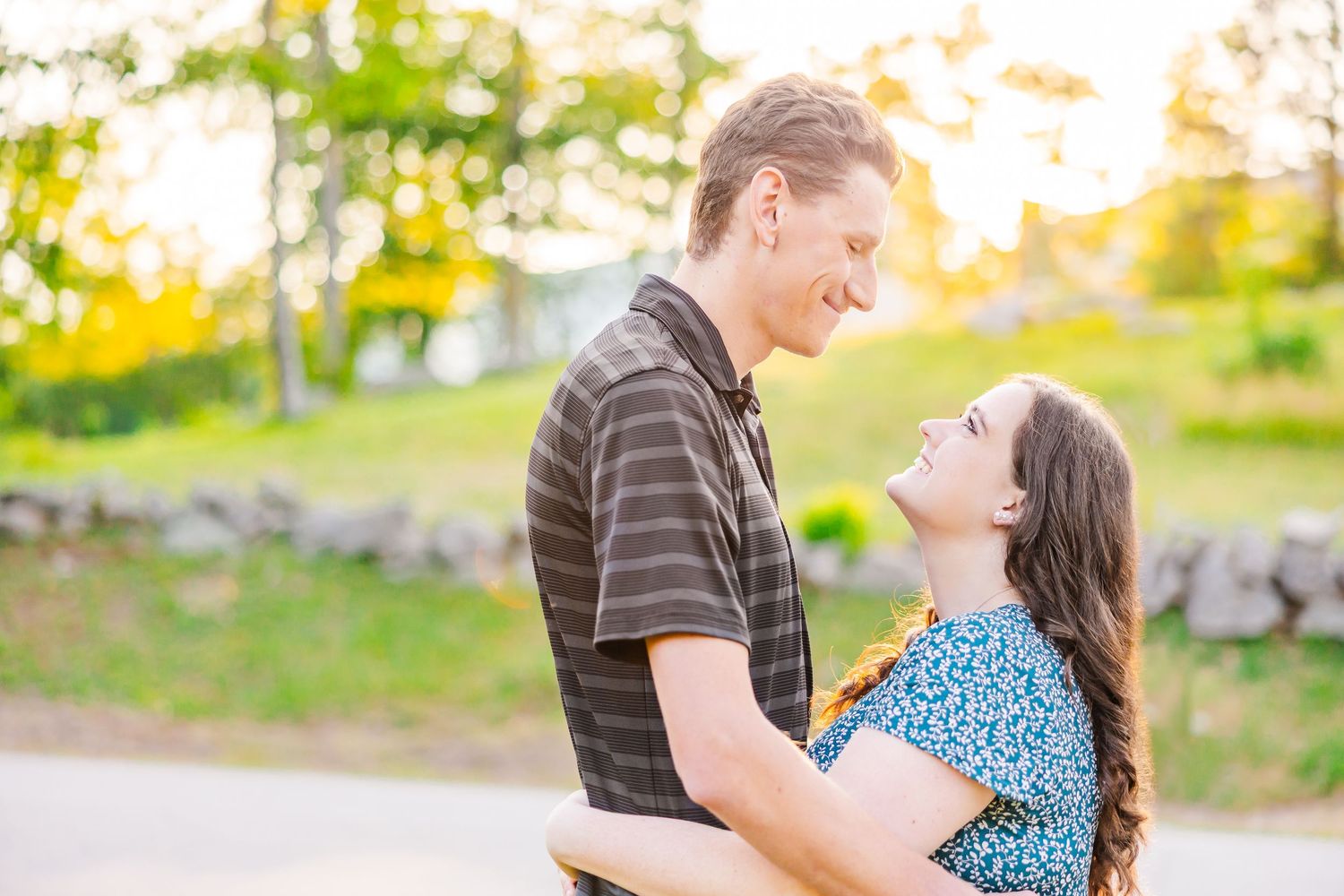 A couple embraces and smiles at each other outdoors on a sunny day with greenery in the background.