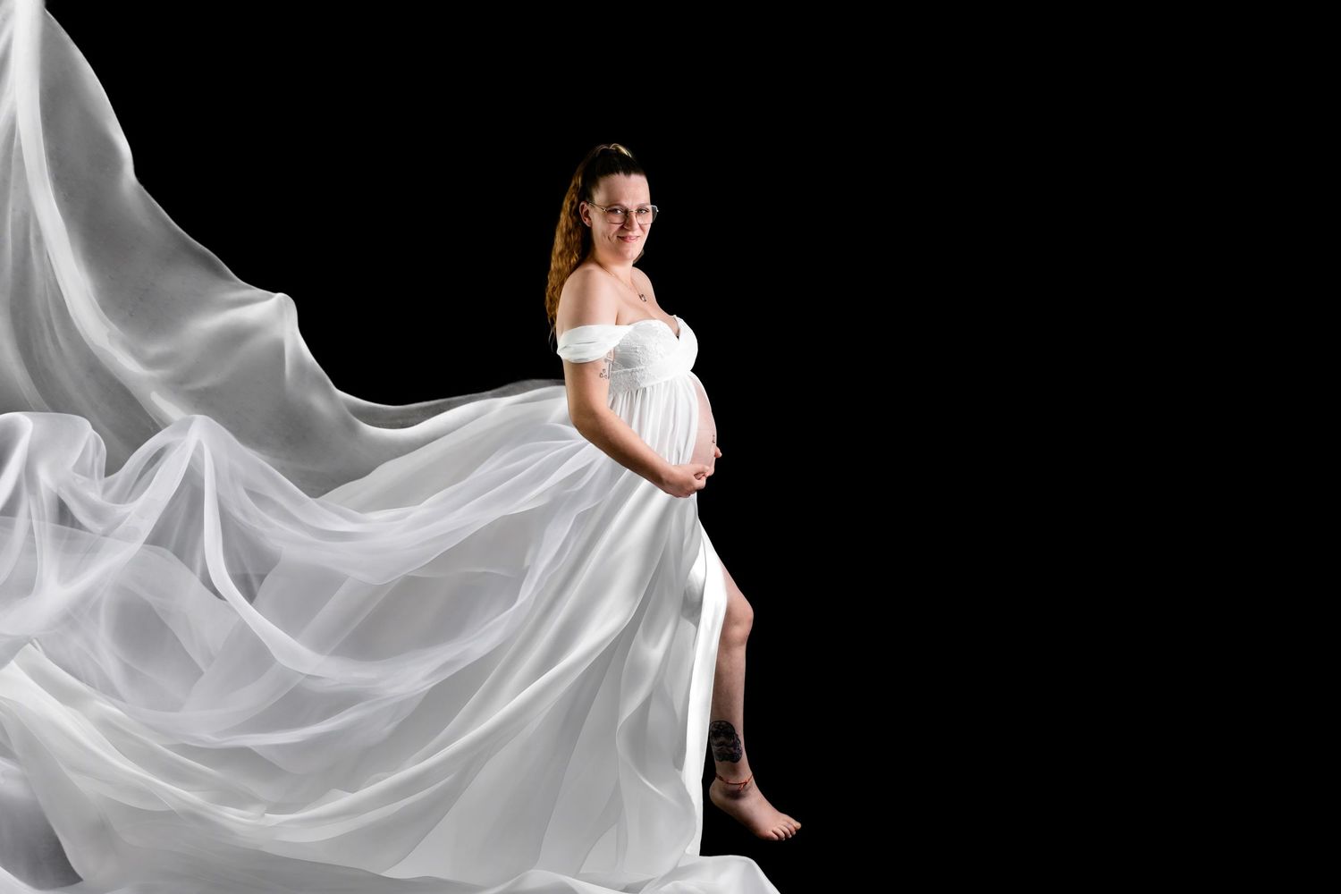 A pregnant woman in a flowing white maternity gown poses elegantly against a black backdrop with billowing fabric.