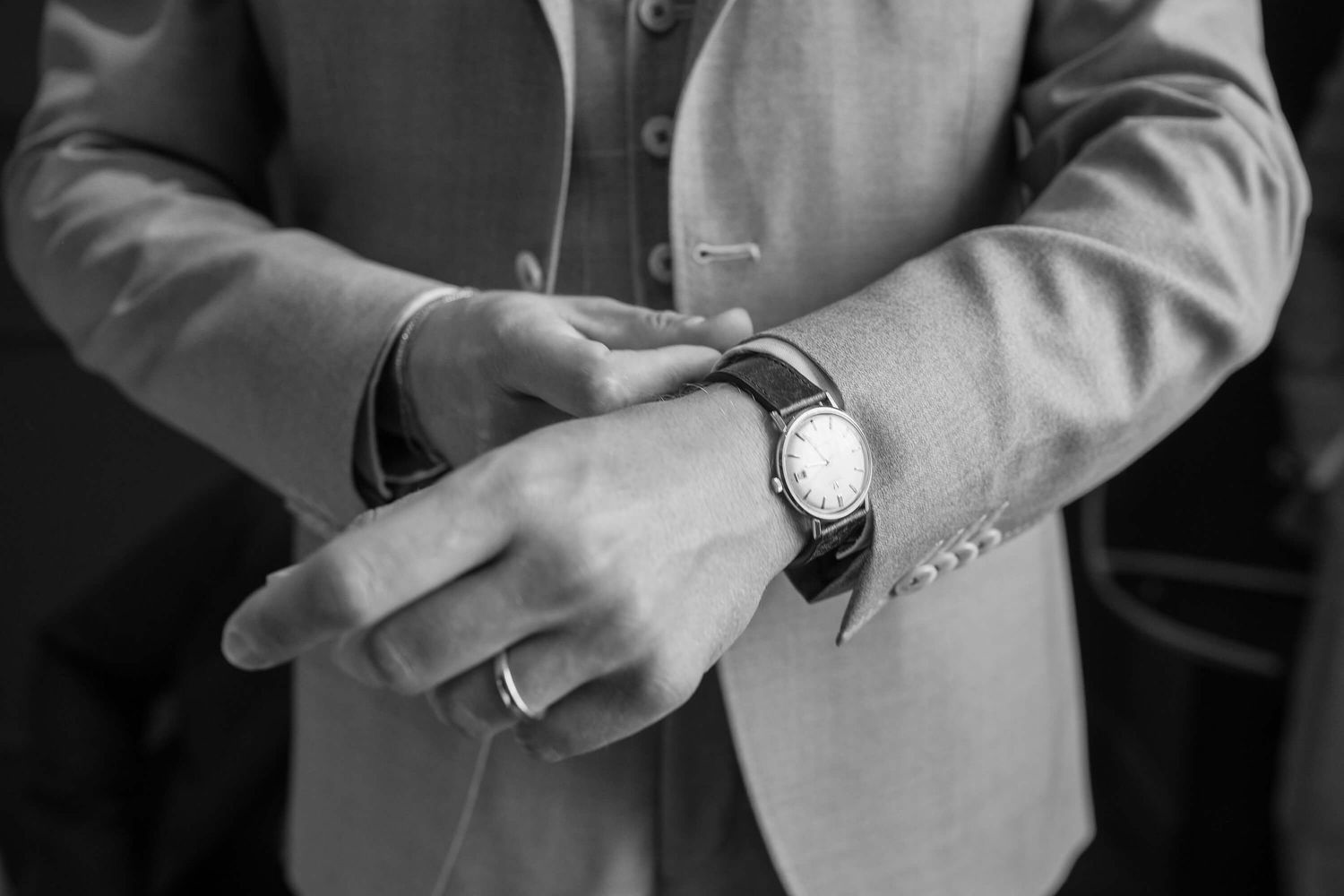 Black and white close-up of hands adjusting a wristwatch while wearing a light colored suit jacket.