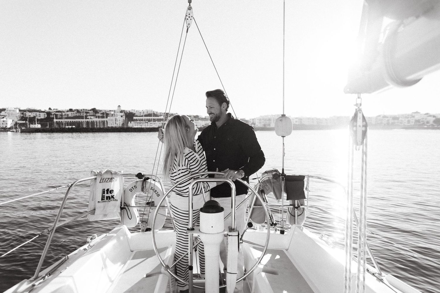 Black and white photo of someone navigating a sailboat on calm waters at sunset with a marina in the background.