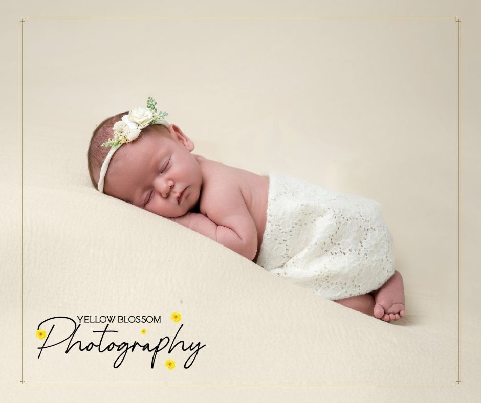 Sleeping newborn swaddled in delicate white lace wrap with tiny floral headband resting peacefully during professional photo shoot.