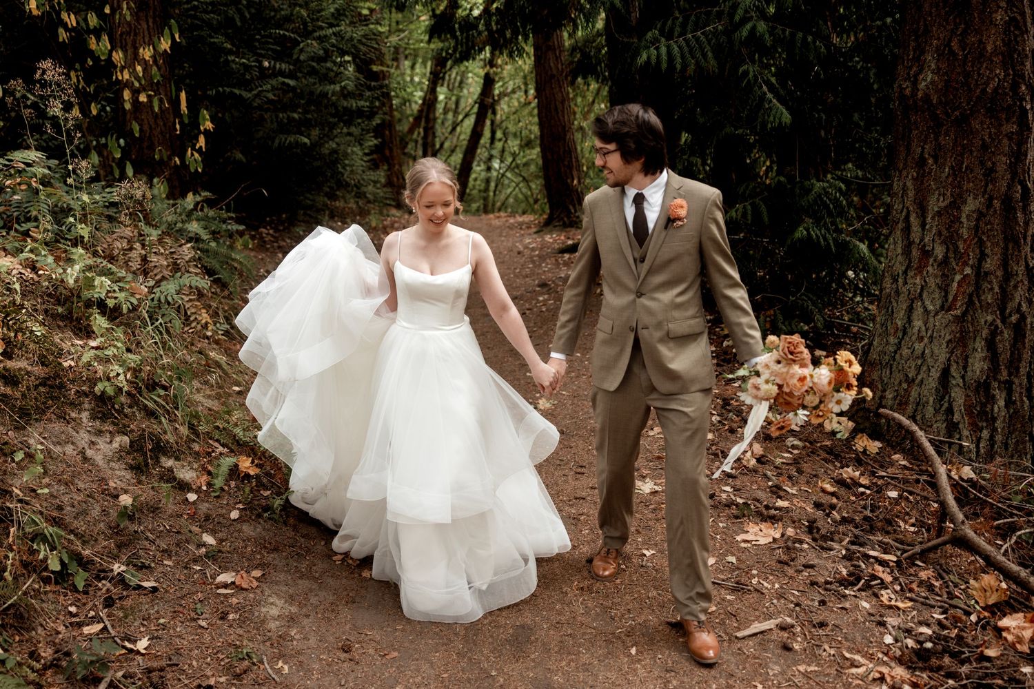 A couple walks hand in hand along a forest path, the bride in a flowing white gown and groom in a tan suit.
