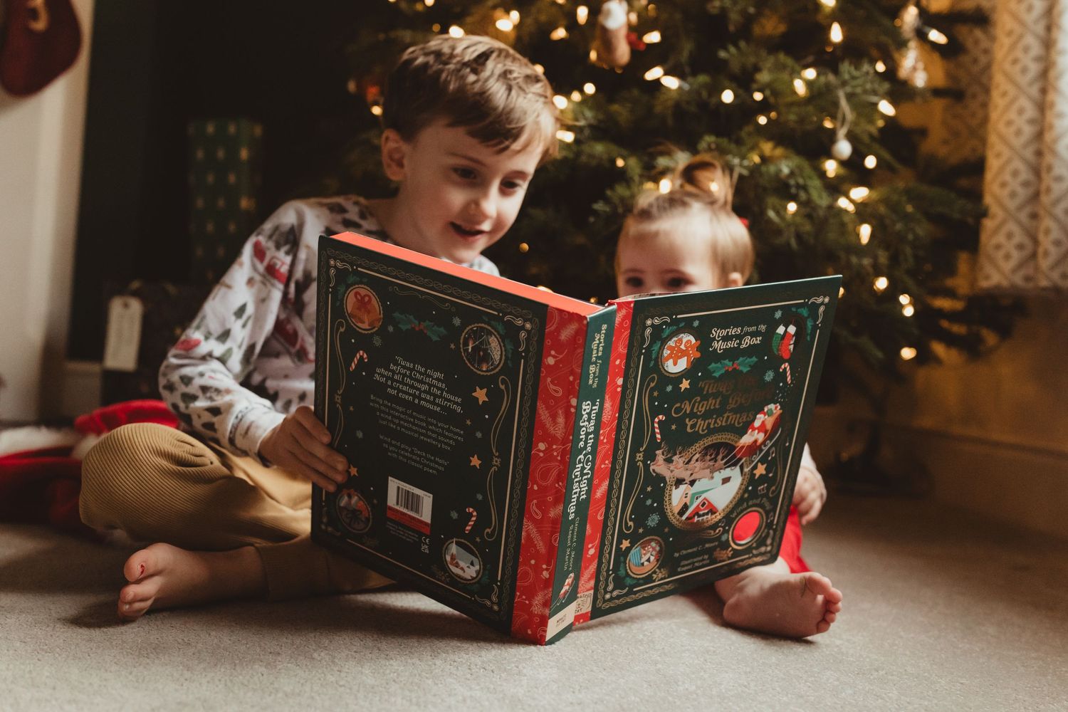 Children sit on floor reading holiday books near a decorated Christmas tree with twinkling lights.