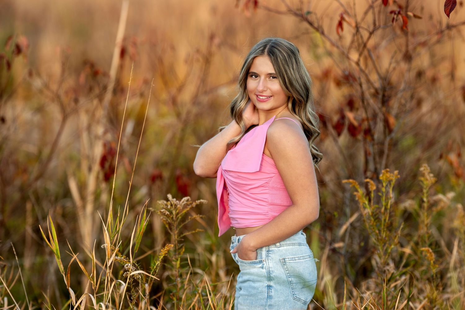 Smiling young woman in pink top and denim shorts poses among autumn wildflowers in golden field.