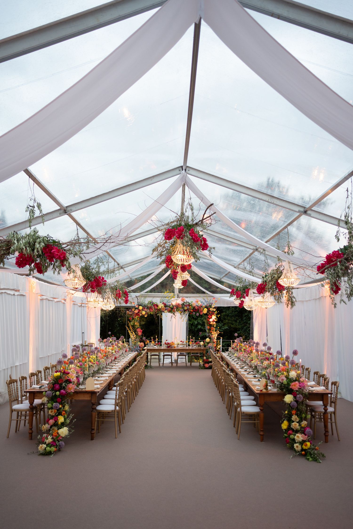 Elegant wedding reception tent with clear ceiling, hanging floral arrangements, and long wooden tables set for dining.