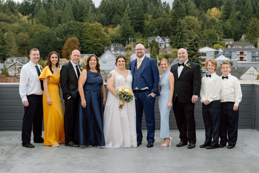 A wedding party in formal attire poses on a rooftop deck with evergreen trees and buildings in the background.