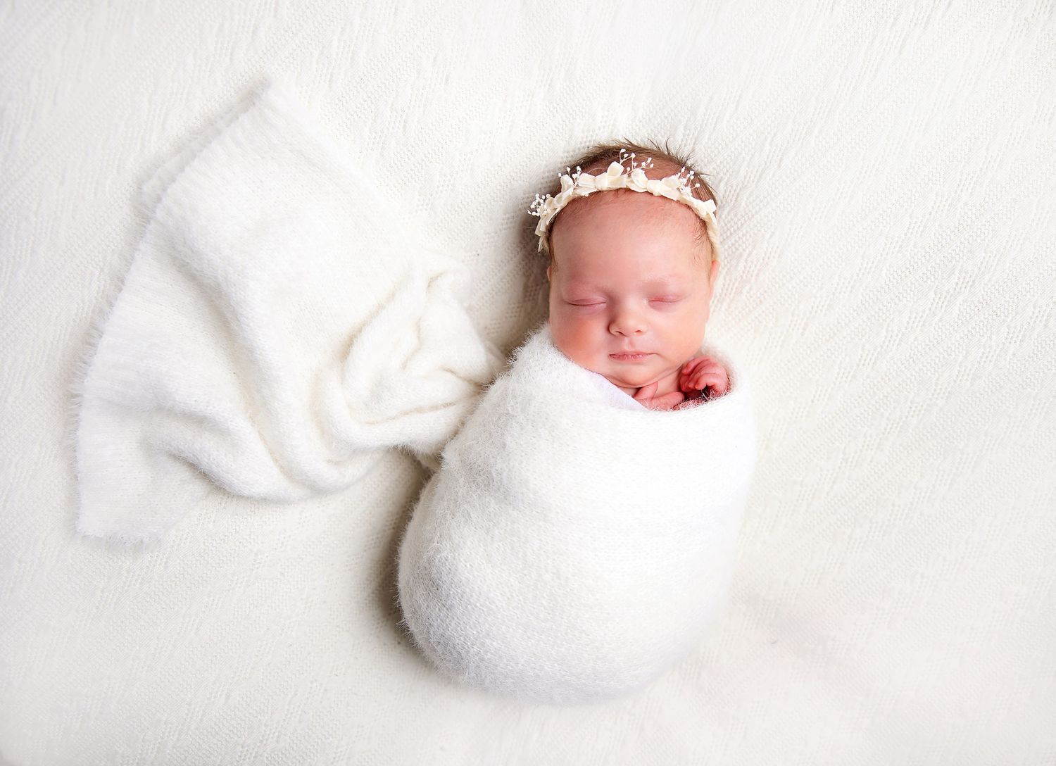 A sleeping newborn swaddled in white fabric with a delicate lace headband laying on soft white background during a London newborn session