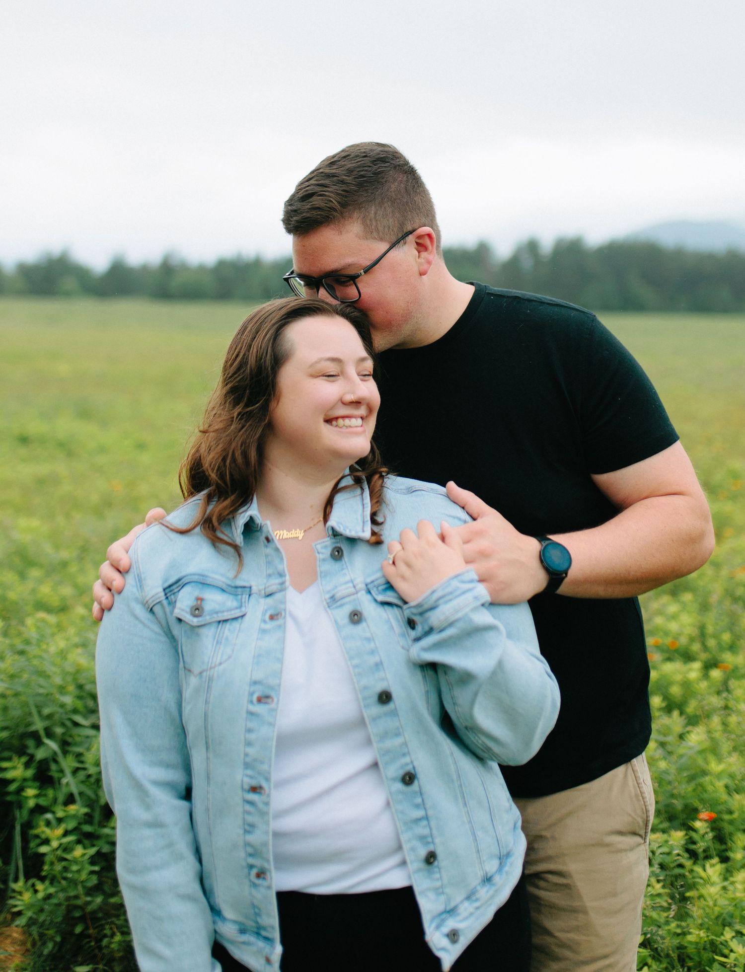 A couple sharing a tender moment together while standing in a sunny meadow with mountains in the background.