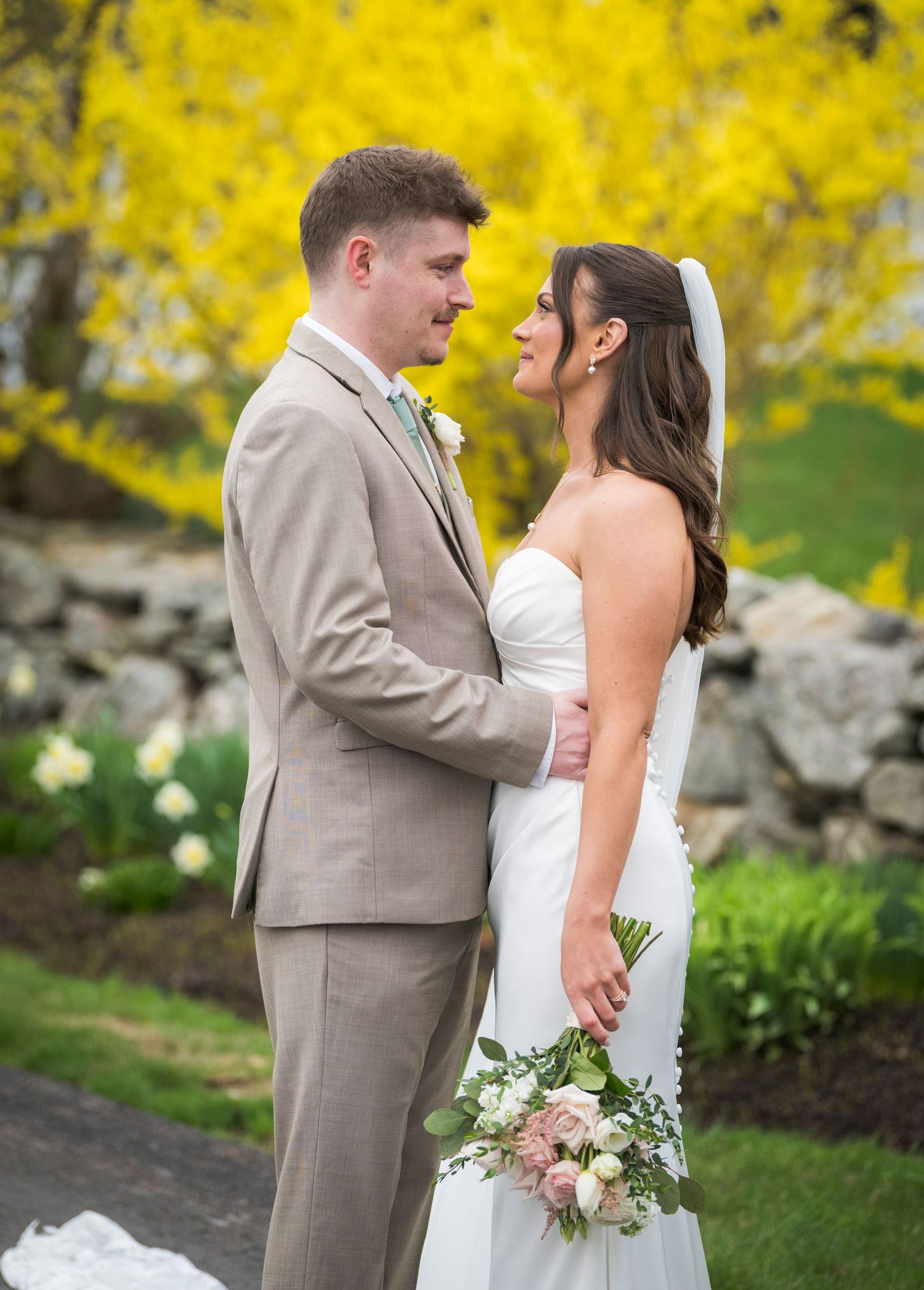 Bride and groom share a romantic moment outdoors in spring, with yellow forsythia blooms and a stone wall backdrop.