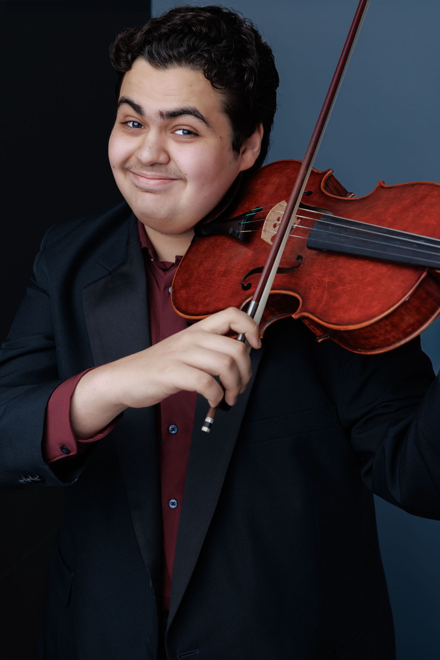 A musician in a black suit jacket poses with a violin against a dark background.