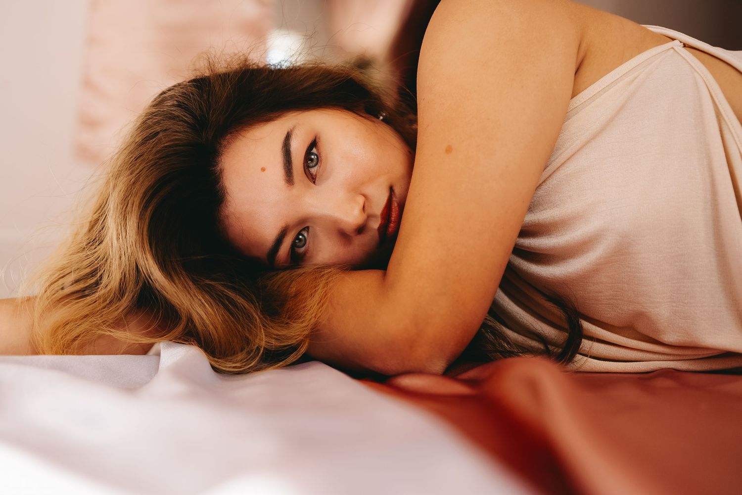 Person with long hair lying on white bedding in soft natural lighting.