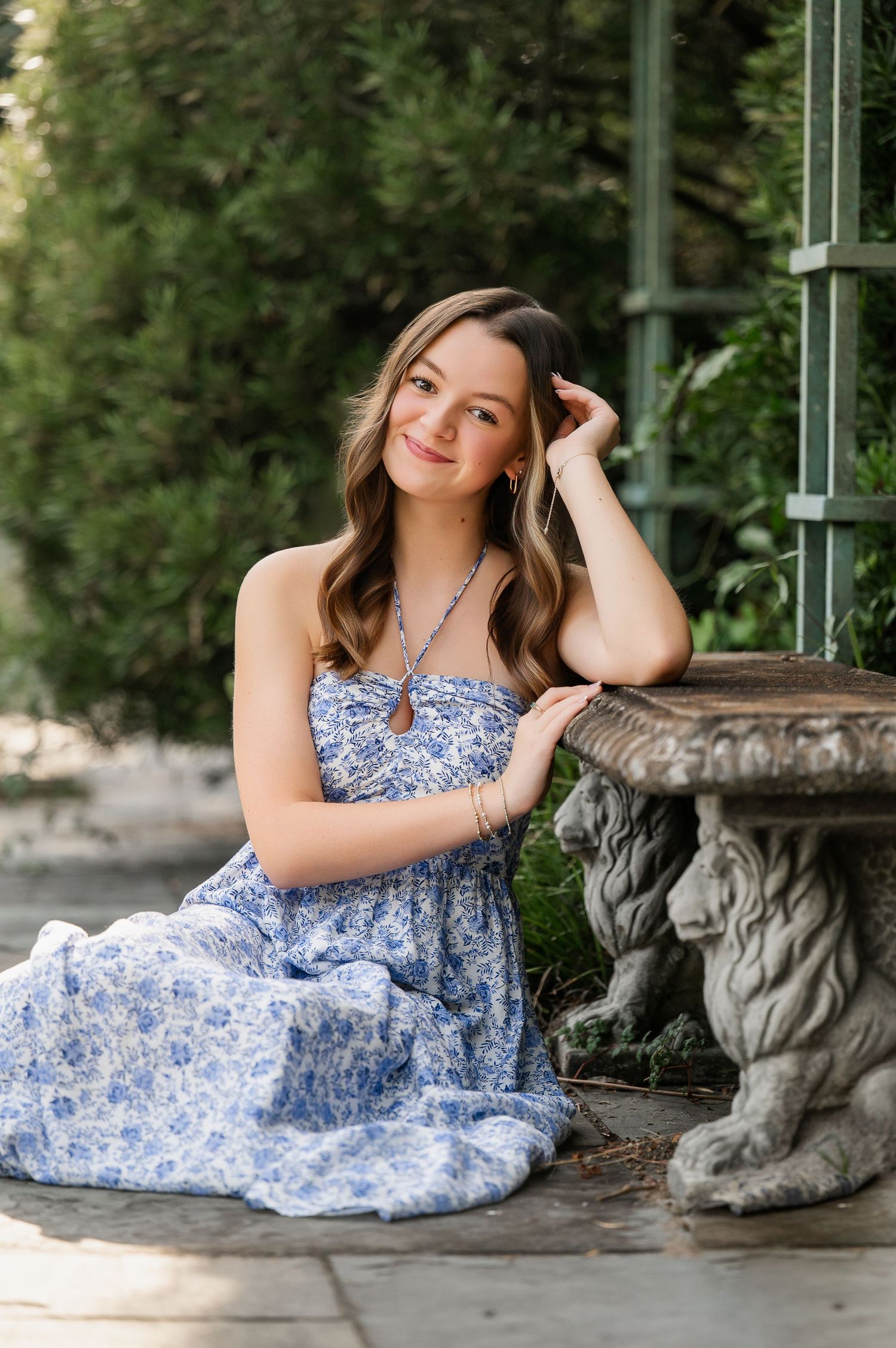 A person in a blue floral dress poses gracefully on a stone bench in a garden setting.