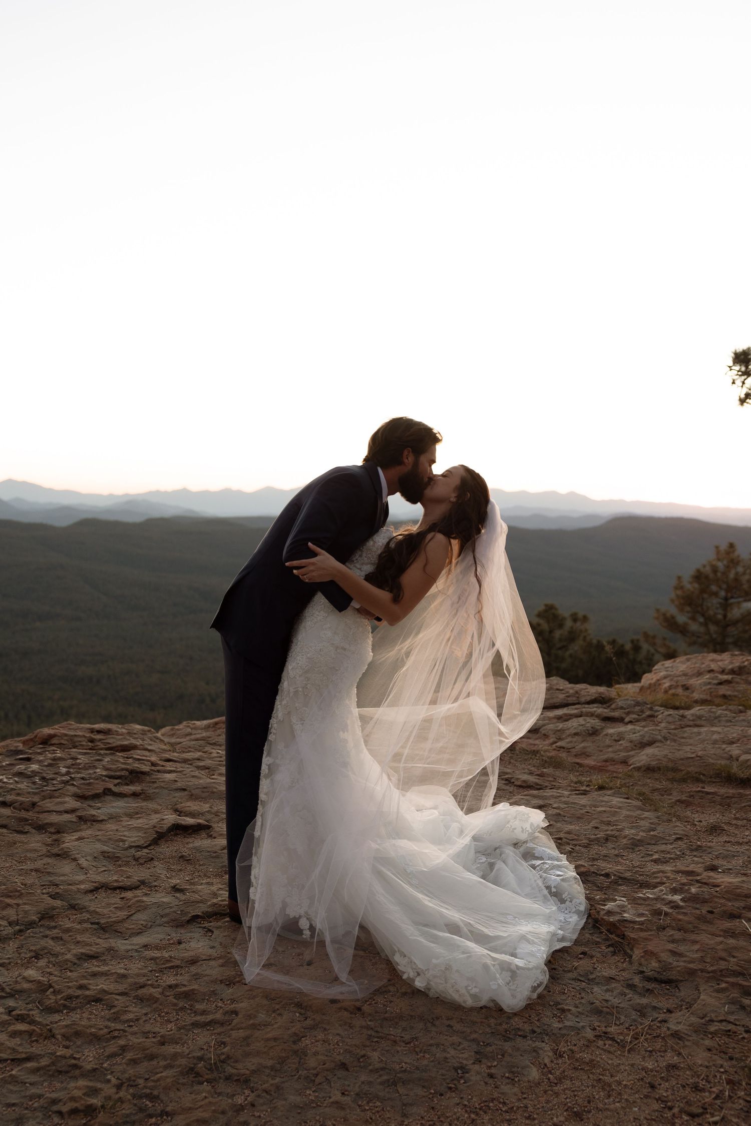 A couple stands together at sunset on a rocky mountain overlook with sweeping views of the landscape below.