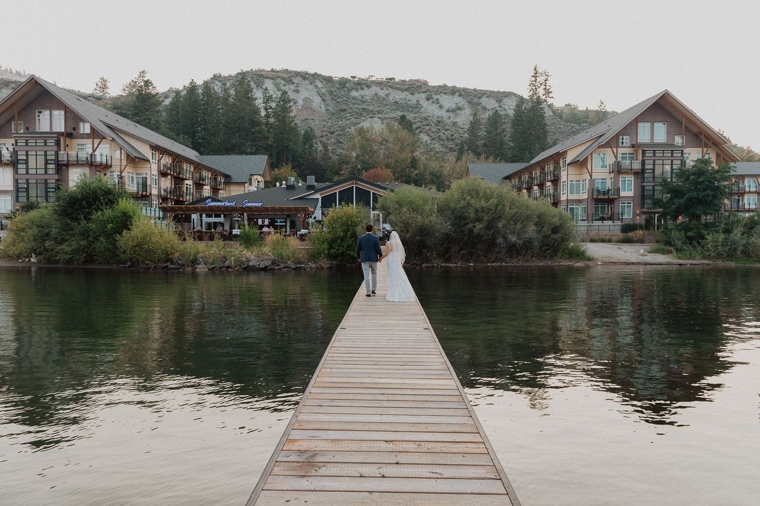 Bride and groom walking hand in hand on wooden dock surrounded by lake and rustic lodges.