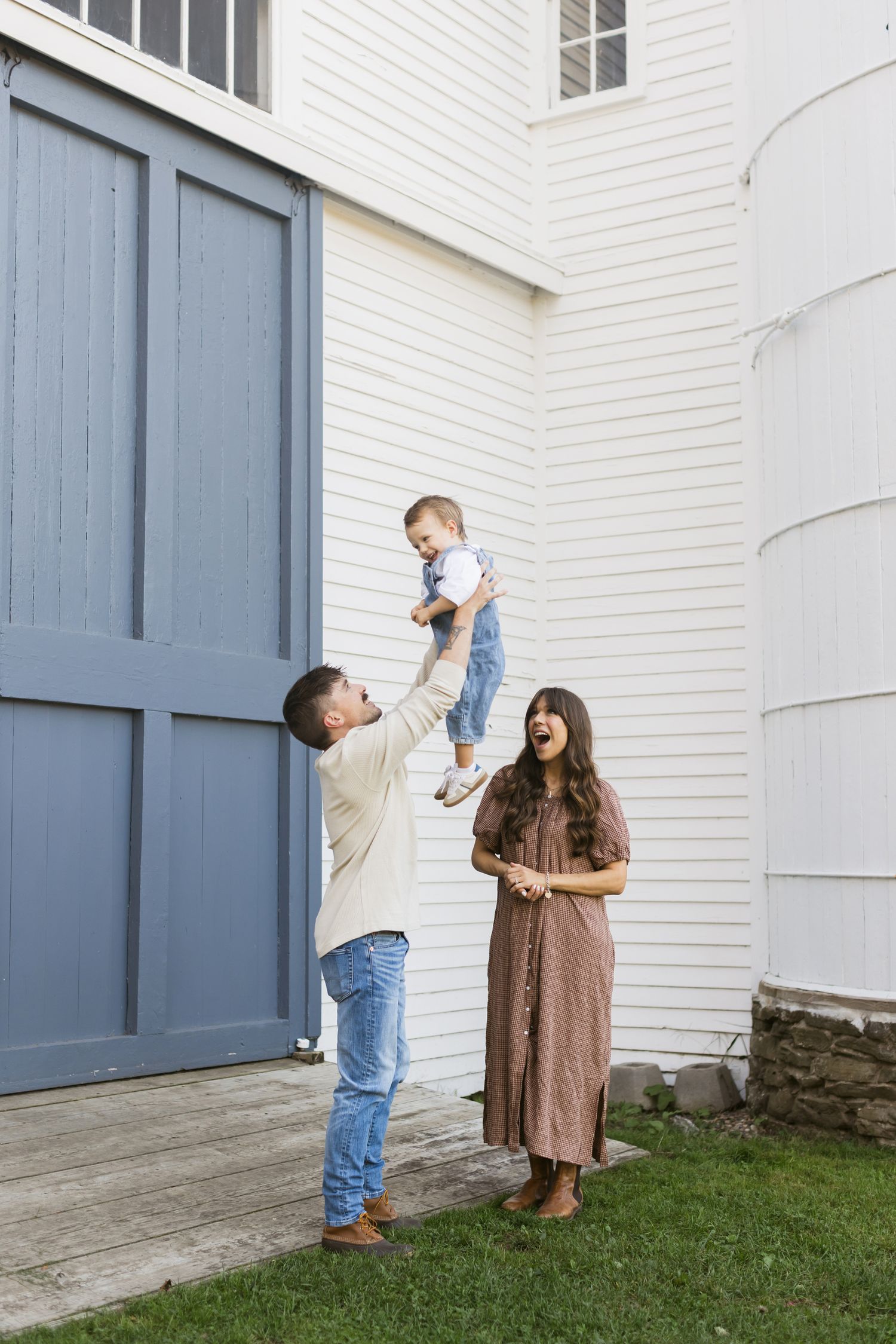 Saltonstall Farm Stratham, New Hampshire family photos.. A mother in a brown gingham dress watches her husband raise their son in the air.